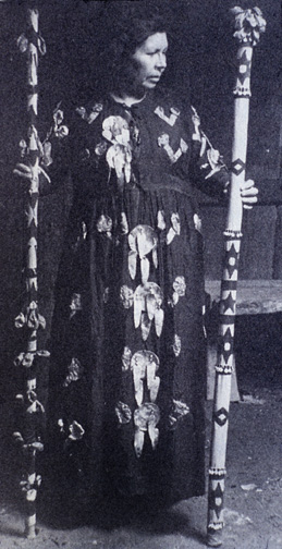 woman in long dress adorned with abalone shells holding decorated wooden staffs