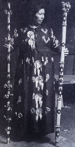 woman in long dress adorned with abalone shells holding decorated wooden staffs